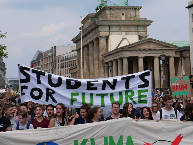 Gruppe von Schülern marschiert in Berlin mit einem leuchtend bunten 'Schüler für die Zukunft'-Schild vor einer Kulisse aus Gebäuden, Bäumen und Himmel.