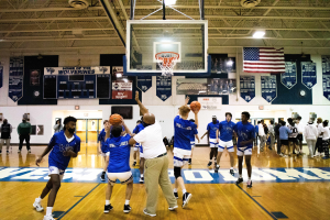 Eine Gruppe von Männern, die Basketball in einer Turnhalle mit einem Korb spielt, Banner, Flaggen, Deckenlampen, ein Projektor und ein sichtbarer Boden.