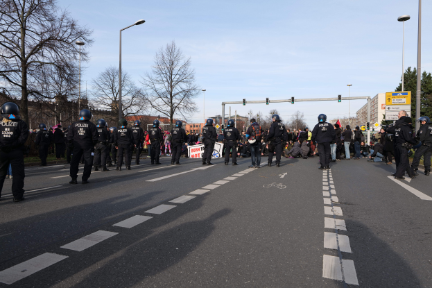 Eine Gruppe von Polizisten in schwarzen Uniformen und Helmen steht am Straßenrand, mit Laternenmasten, Ampeln, Bäumen, Gebäuden und einem klaren blauen Himmel im Hintergrund.