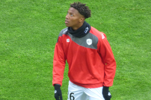 Ein junger Mann in einer roten Jacke und weißen Shorts steht auf einem Fußballfeld und schaut rechts mit entschlossener Miene, wobei er Baseballhandschuhe trägt.