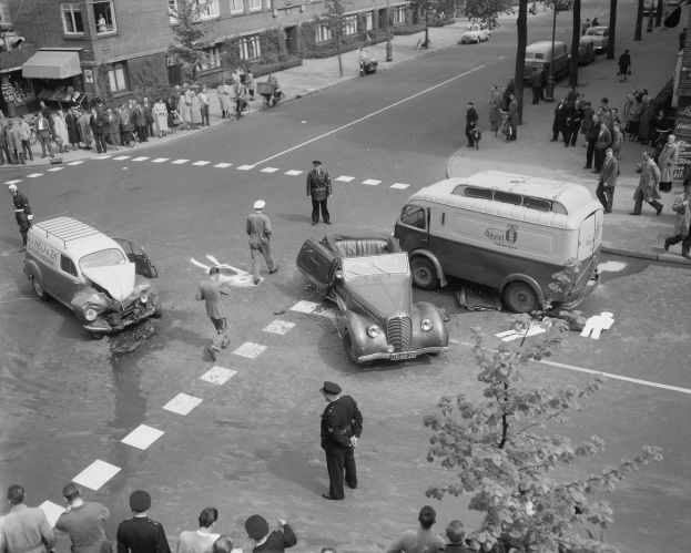 Schwarz-weiß-Szene eines Autounfalls an einer Straßenkreuzung mit mehreren Fahrzeugen, darunter ein Van, und einer Gruppe von Schaulustigen; Gebäude, Bäume, Pfähle und Texttafeln sind im Hintergrund zu sehen.