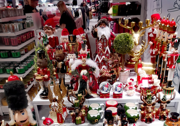 Ladeninneres weihnachtlich dekoriert mit Tischen, die Spielzeug, Tassen und andere Gegenstände zeigen, Regalen mit Waren und Menschen im Hintergrund unter festlicher Beleuchtung.