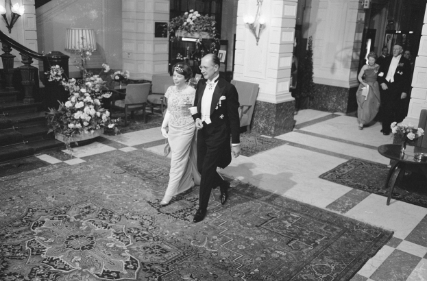 Schwarz-weißes Foto einer Braut und eines Bräutigams, die durch die Lobby eines Hotels gehen, umgeben von einer Gruppe von Menschen, die auf dem Boden stehen, einem Tisch mit einer Blumenvase darauf, einigen Stühlen, Blumen in Töpfen, Lampen, einer Treppe, einem Geländer und einer Wand.