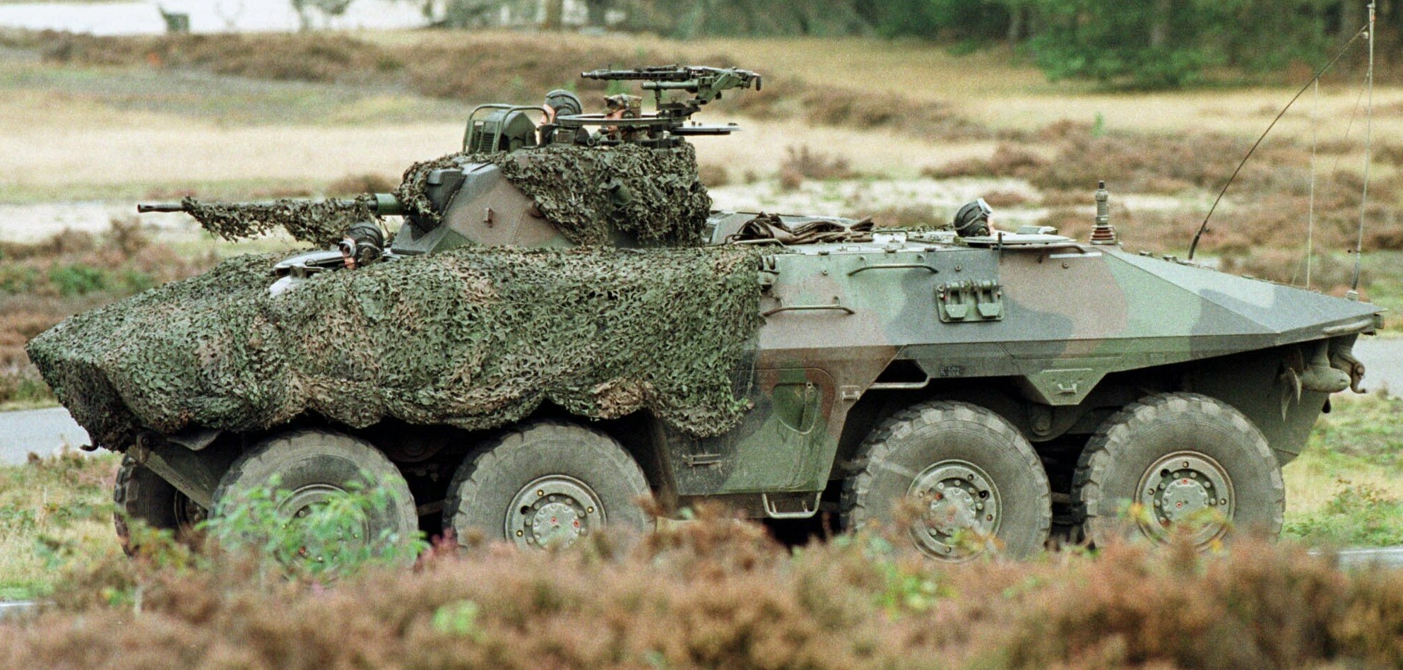 Militärisches Personalfahrzeug mit montierter großer Kanone, das auf einer Straße neben einem Wald fährt.