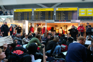 Eine große Gruppe von Menschen sitzt und steht in einem Flughafen während einer Protestaktion, mit Informationsschildern, Schaufensterpuppen in Kleidern und Deckenleuchten im Hintergrund.