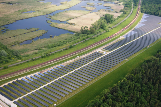 Luftaufnahme einer großen Solarfarms in einem Feld mit Solarpanelen, umgeben von Bäumen, Gras, Pflanzen und Wasser, mit einem Zug auf einer nahen Eisenbahnschiene.