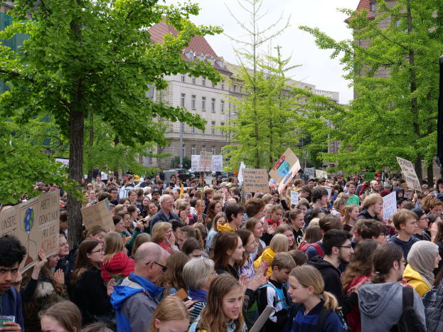 Eine große Menge von Menschen protestiert vor einem Gebäude in Berlin, mit Schildern und einem Lautsprecher auf der rechten Seite.