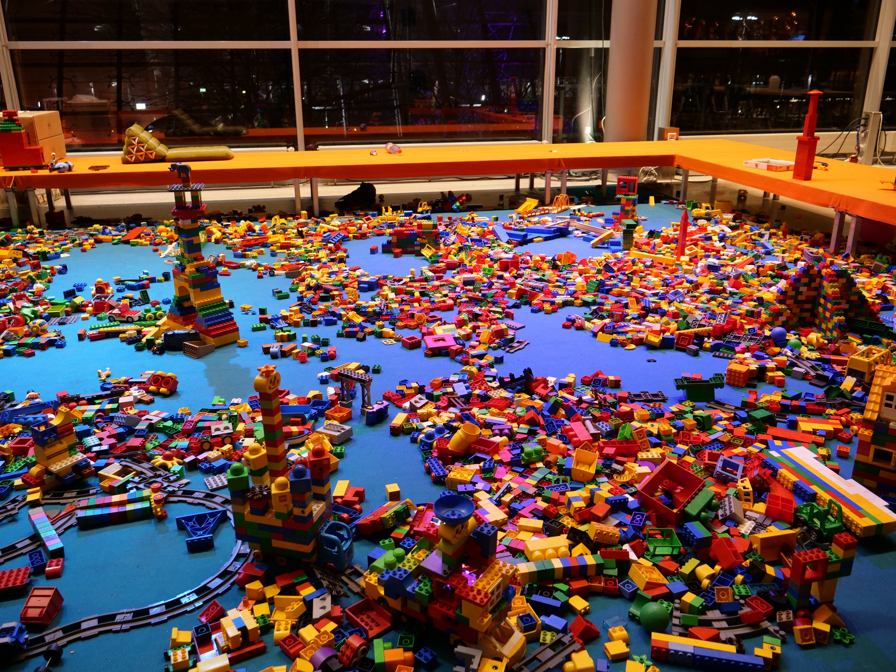 Ein geräumiger Raum mit farbenfrohen LEGO-Bausteinen, die auf dem Boden verstreut sind, umgeben von Tischen und Säulen, mit einem Blick durch Glasfenster auf Menschen und beleuchtete Ausstellungen.