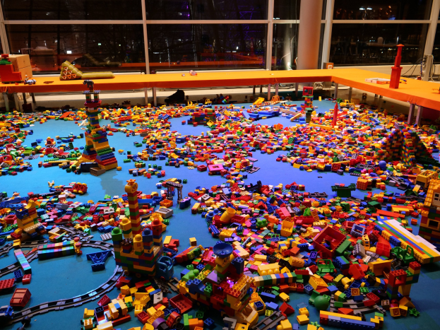 Ein geräumiger Raum mit farbenfrohen LEGO-Bausteinen, die auf dem Boden verstreut sind, umgeben von Tischen und Säulen, mit einem Blick durch Glasfenster auf Menschen und beleuchtete Ausstellungen.