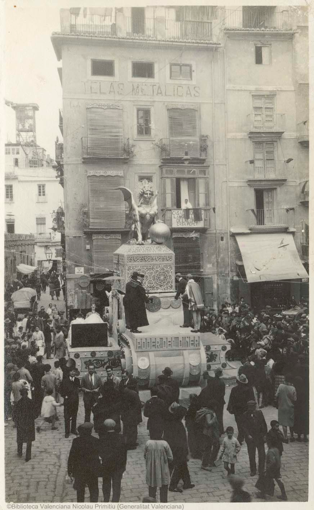 Ein altes Schwarz-Wei├č-Foto eines Umzugs mit einer Menge um einen geschm├╝ckten Wagen mit einer Statue, umgeben von Geb├Ąuden mit Fenstern und Balkonen, und Text unten.
