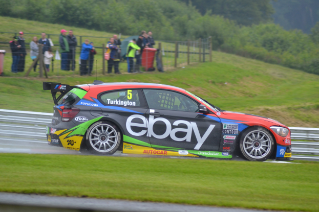 Ein Auto auf einer Rennstrecke mit Rauch, umgeben von Gras, einem Zaun, Mülltonnen, Menschen, Bäumen und einem klaren blauen Himmel.