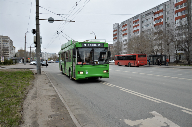 Ein grüner Bus fährt eine Straße entlang, die von hohen Gebäuden gesäumt ist, mit Gras, Strommasten und Drähten auf der linken Seite, Bäumen im Hintergrund und einem klaren blauen Himmel darüber.