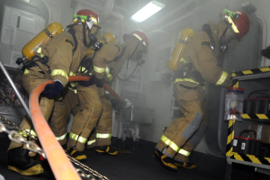Feuerwehrleute in Schutzausrüstung arbeiten an einem Löschwagen mit Equipment-Regalen und sichtbarem Hintergrund-Infrastruktur.