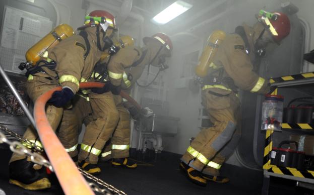 Feuerwehrleute in Schutzausrüstung arbeiten an einem Löschwagen mit Equipment-Regalen und sichtbarem Hintergrund-Infrastruktur.