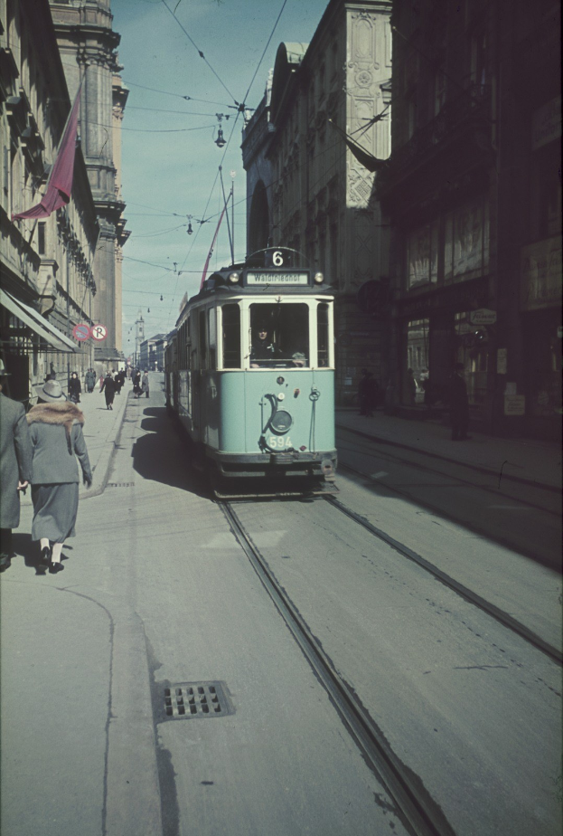 Altes Schwarz-Weiß-Foto einer Straßenbahn, die auf einer Stadtstraße mit Fußgängern auf dem linken Gehweg, Gebäuden und Strommasten im Hintergrund unter einem klaren blauen Himmel fährt.