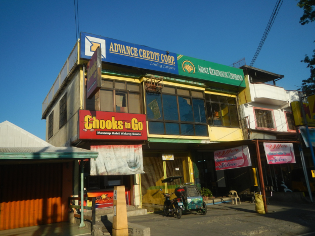 Ein Gebäude mit einer Tafel, auf der "Advance Credit Corp" steht, auf einer Straße mit Fahrzeugen, Werbetafeln, Strommasten und Bäumen unter einem klaren blauen Himmel.