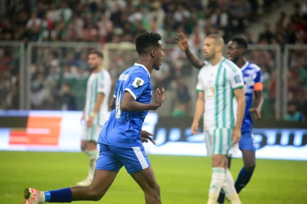 Ein Fussballspieler in einem blauen und weissen Trikot feiert mit erhobenen Armen, umgeben von einer Gruppe von Menschen in weissen und grünen Trikots, mit einem unscharfen Hintergrund, der ein weisses und blaues Brett und ein Geländer zeigt.