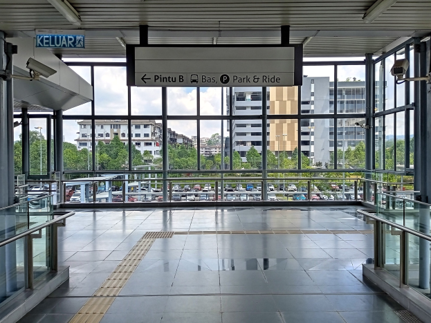 Leeres Parkhaus mit Glaswänden und Geländern, ein Schild, Deckenleuchten und eine Außenansicht von Fahrzeugen, Bäumen, Gebäuden und einem bewölkten Himmel.
