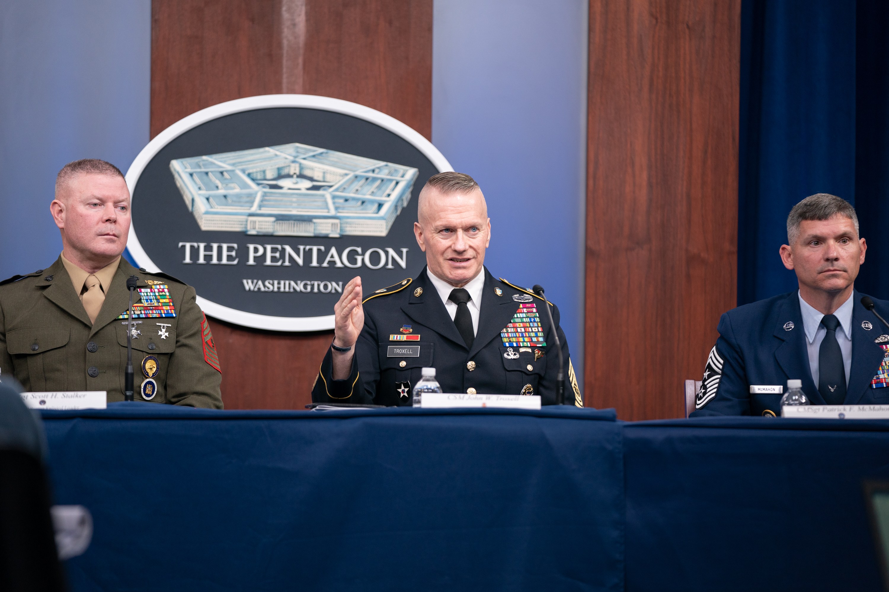 Drei Männer in Militäruniformen sitzen an einem Tisch mit Namensschildern, Flaschen, Mikrofonen und einem blauen Tuch und diskutieren mit einem Verteidigungsminister im Hintergrund.