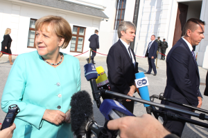 Bundeskanzlerin Angela Merkel spricht mit Reportern vor dem Kanzleramt in Berlin, umgeben von Menschen mit Mikrofonen, mit Gebäuden im Hintergrund.