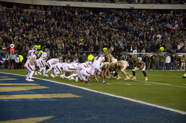 Ein Fussballspiel in einem Stadion mit Spielern in Sportkleidung und Helmen auf dem Feld, Zuschauern, Gel"andern und Tafeln mit Text im Hintergrund.