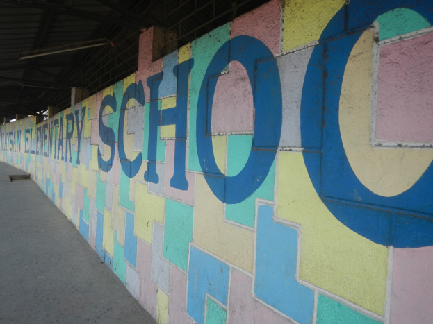 Buntes Wandgemälde an einer Gebäudeseite mit dem Wort "Schule" in leuchtenden Blautönen, Grün, Gelb und Rot, beleuchtet von Deckenlampen.