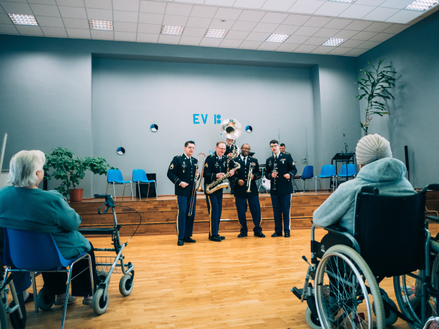 Eine Gruppe von Menschen im Rollstuhl, die in einem Raum mit Stühlen, Pflanzen und Deckenleuchten Musikinstrumente spielen, während einige Individualsitzend zuschauen.