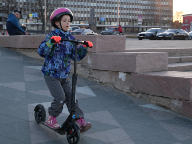Ein junger Junge mit Helm und Handschuhen fährt einen Roller eine Treppe hinunter, vorbei an Fahrzeugen, Menschen, Bäumen, Pfosten, Brettern, Gebäuden und einem klaren blauen Himmel.