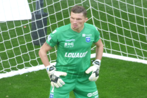 Ein Torwart in grüner Uniform steht vor einem Fußballtor auf saftig grünem Rasen, trägt Handschuhe.