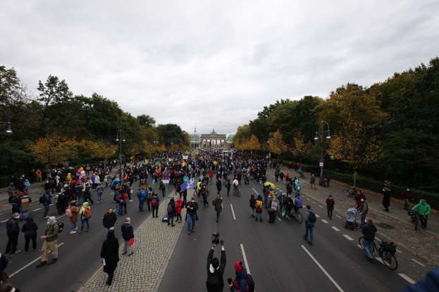 Eine große Gruppe von Menschen marschiert auf einer von Bäumen gesäumten Straße in Berlin, mit Kameras ausgerüstet, mit einem Gebäude und einem klaren Himmel im Hintergrund.