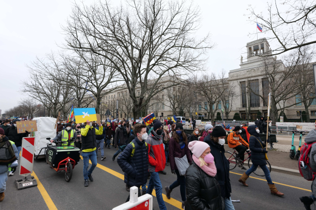 Eine große Gruppe von Menschen nimmt an einer Protestdemonstration auf einer Straße in Washington, D.C. teil, einige halten Schilder und Banner, andere fahren Fahrräder, und Schilder, Bäume und ein klarer blauer Himmel im Hintergrund.
