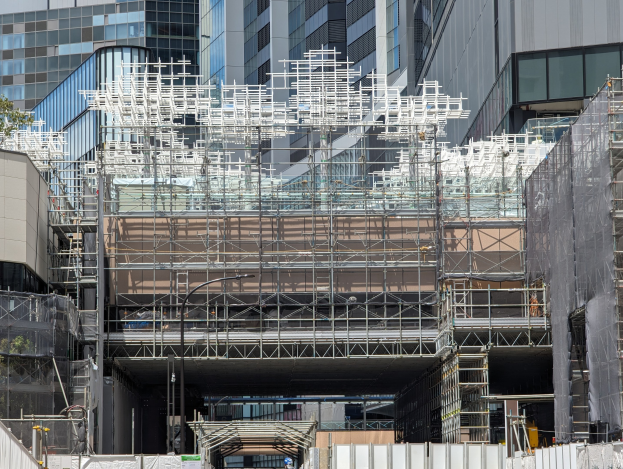 Großes Gebäude mit Gerüst, das renoviert wird, umgeben von Bäumen links und einer Straße unten in einer Stadtkulisse.