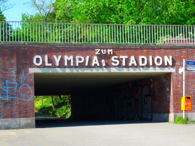 Der Eingang zum Olympiastadion in Berlin, Deutschland, mit einer Brücke, einem Metallzaun, einer Tafel, einem Kasten, einigen Pflanzen, Gras, einer Baumgruppe und einem bewölkten Himmel.