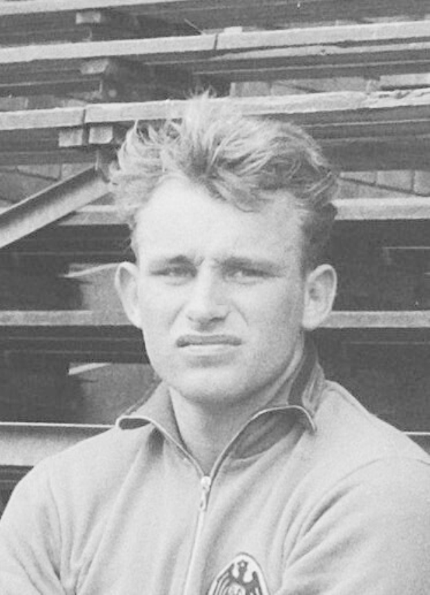 Schwarzes und weißes Foto eines Mannes in einer Jacke vor einem Haufen Holzplanken.