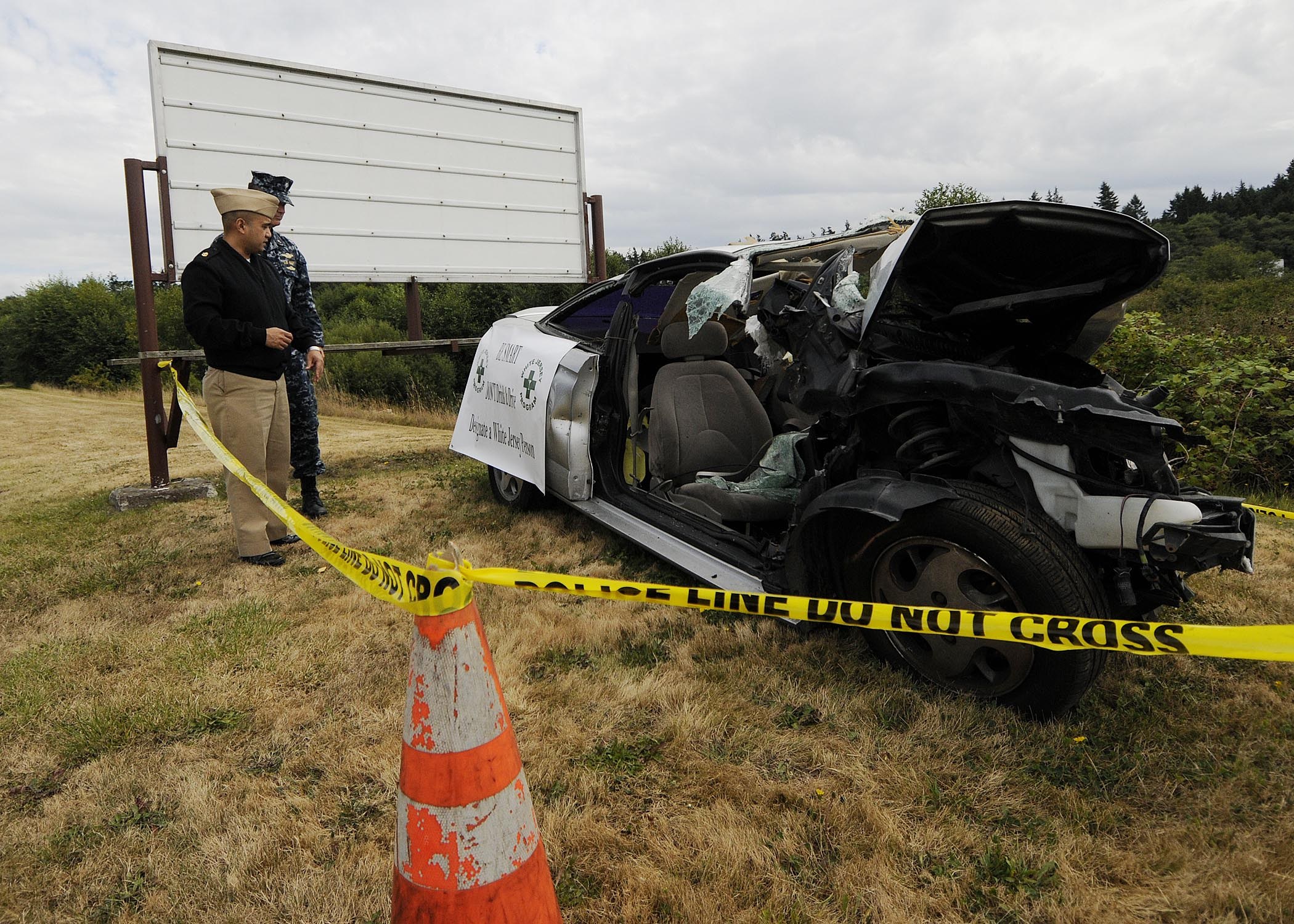 Ein Polizist in Uniform steht neben einem schwer beschädigten Auto am Straßenrand mit eingedrückter Haube und zersplitterten Fenstern, bei einem Verkehrskegel und einem bandagierten Pfahl, mit Bäumen und einem bewölktem Himmel.