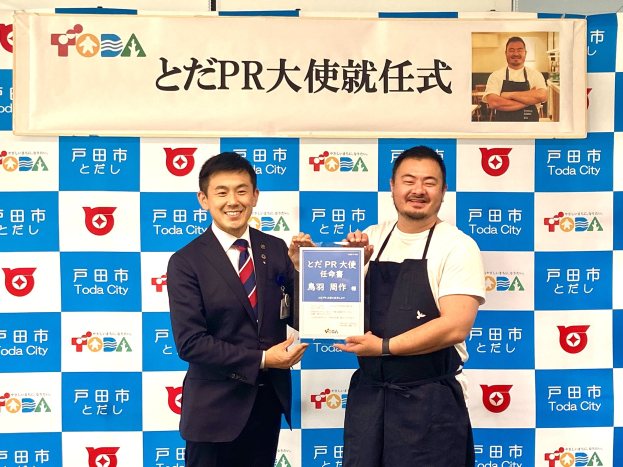 Zwei Männer mit einem Zertifikat, die strahlen und ein "Toda City Wins PR Award"-Banner sowie ein Personenfoto im Hintergrund halten.