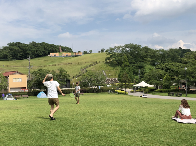 Menschen spielen Badminton in einem Park mit Zelten und Gebäuden im Hintergrund.