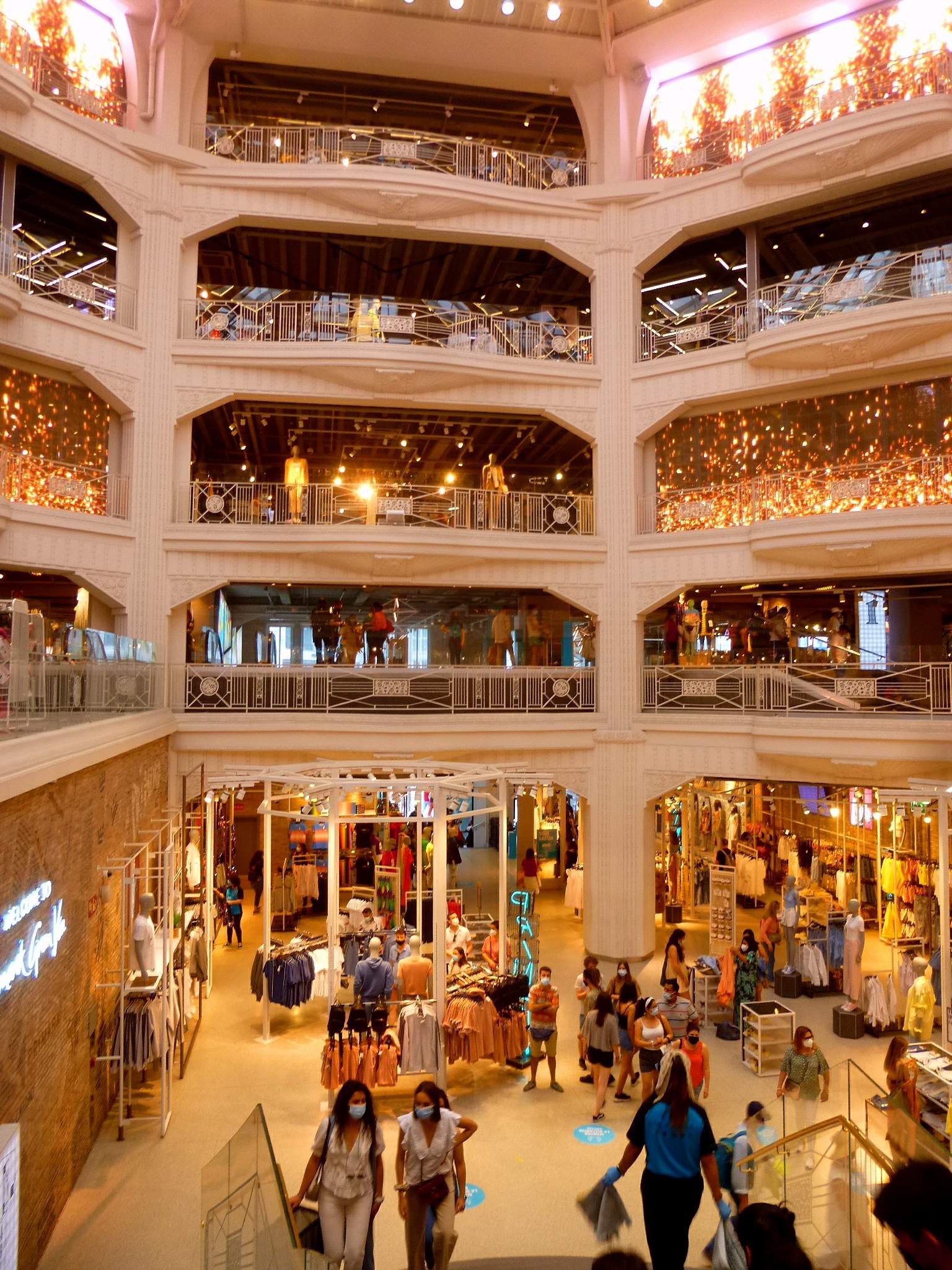 Ein belebtes Einkaufszentrum mit Menschen, die mit Einkaufstaschen und Schaufensterpuppen in Kleidern umhergehen, umgeben von Geländern, Lampen und Wandtafeln.