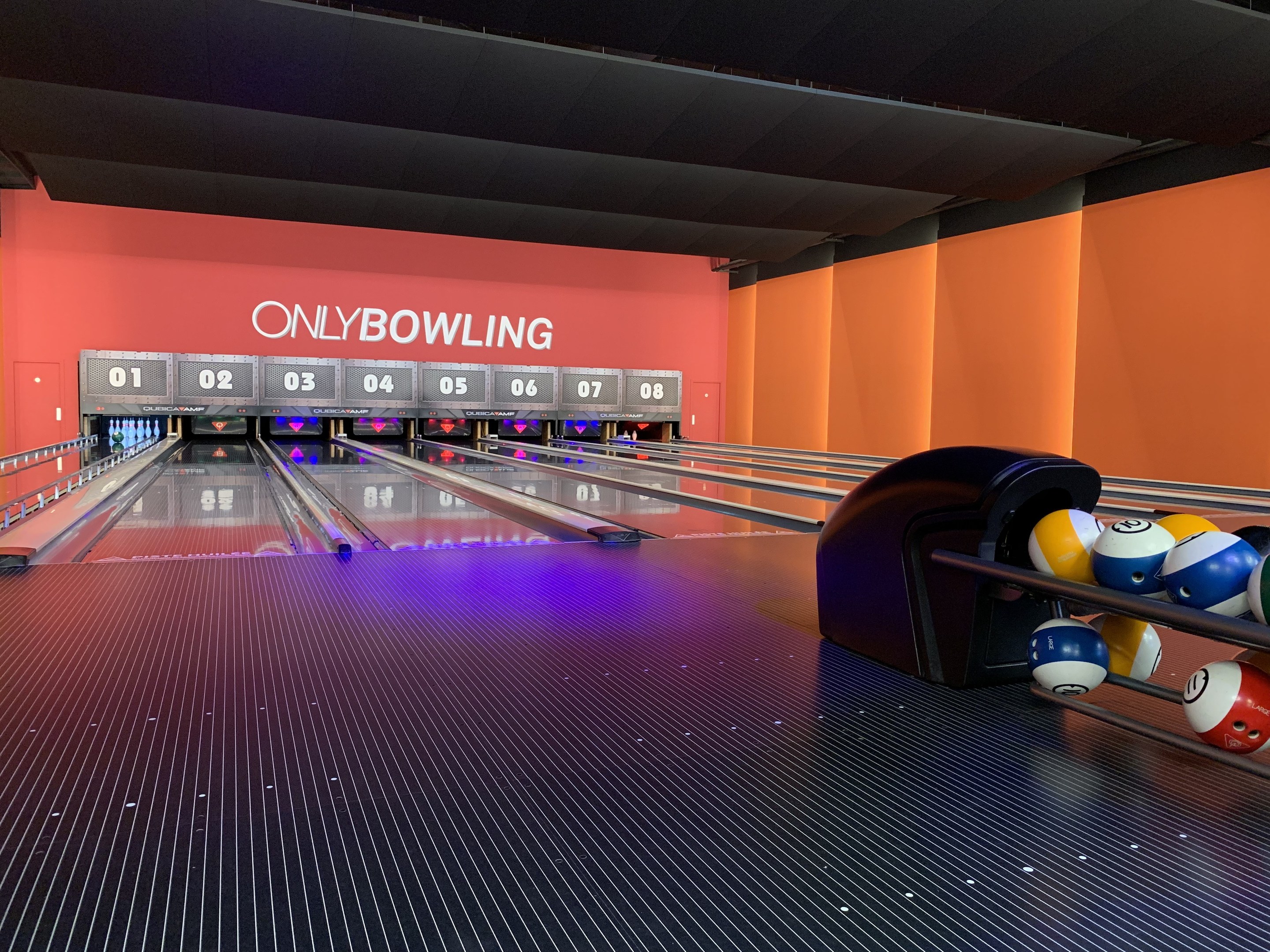 Eine Bowlingbahn mit Bowlingkugeln rechts, Pins in der Mitte, einer textbedeckten Wand im Hintergrund und einer sichtbaren Decke.