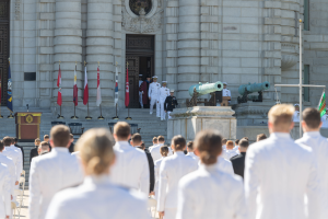 Eine Gruppe von Menschen in weißen Uniformen und Mützen steht in Formation vor einem Gebäude mit Säulen und einer Tür, mit Fahnen, einem Podium mit Mikrofon, Stufen und Kanonen im Hintergrund.