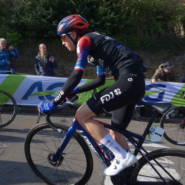 Professionelle Radrennfahrerin Katie Archibald trägt einen Helm, eine Brille, Handschuhe und Schuhe und fährt ein Fahrrad auf einer Straße neben einer Menschenmenge, mit einem Banner, einer Wand, Bäumen und einem Gebäude im Hintergrund.