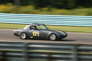 Ein Auto mit einer Person mit Helm darin fährt auf einer Straße mit Absperrungen auf beiden Seiten und Bäumen im Hintergrund.