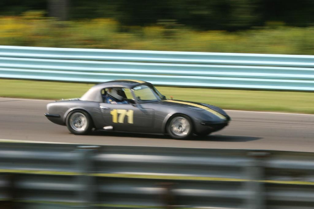 Ein Auto mit einer Person mit Helm darin fährt auf einer Straße mit Absperrungen auf beiden Seiten und Bäumen im Hintergrund.
