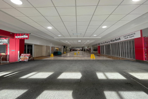 Innenansicht eines Einkaufszentrums mit Deckenleuchten, einem Stuhl und einer Tafel, die links mit "Sydbury Market - Vorher und Nachher" beschriftet ist, sowie verstreuten Gegenständen auf der rechten Seite und auf dem Boden.