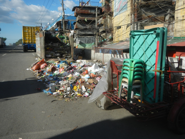 Ein Lastwagen neben einem Haufen Müll auf einer Straße, mit einem Wagen mit Plastikstühlen rechts daneben und Gebäuden, Strommasten, Bäumen und einem bewölkten Himmel im Hintergrund.