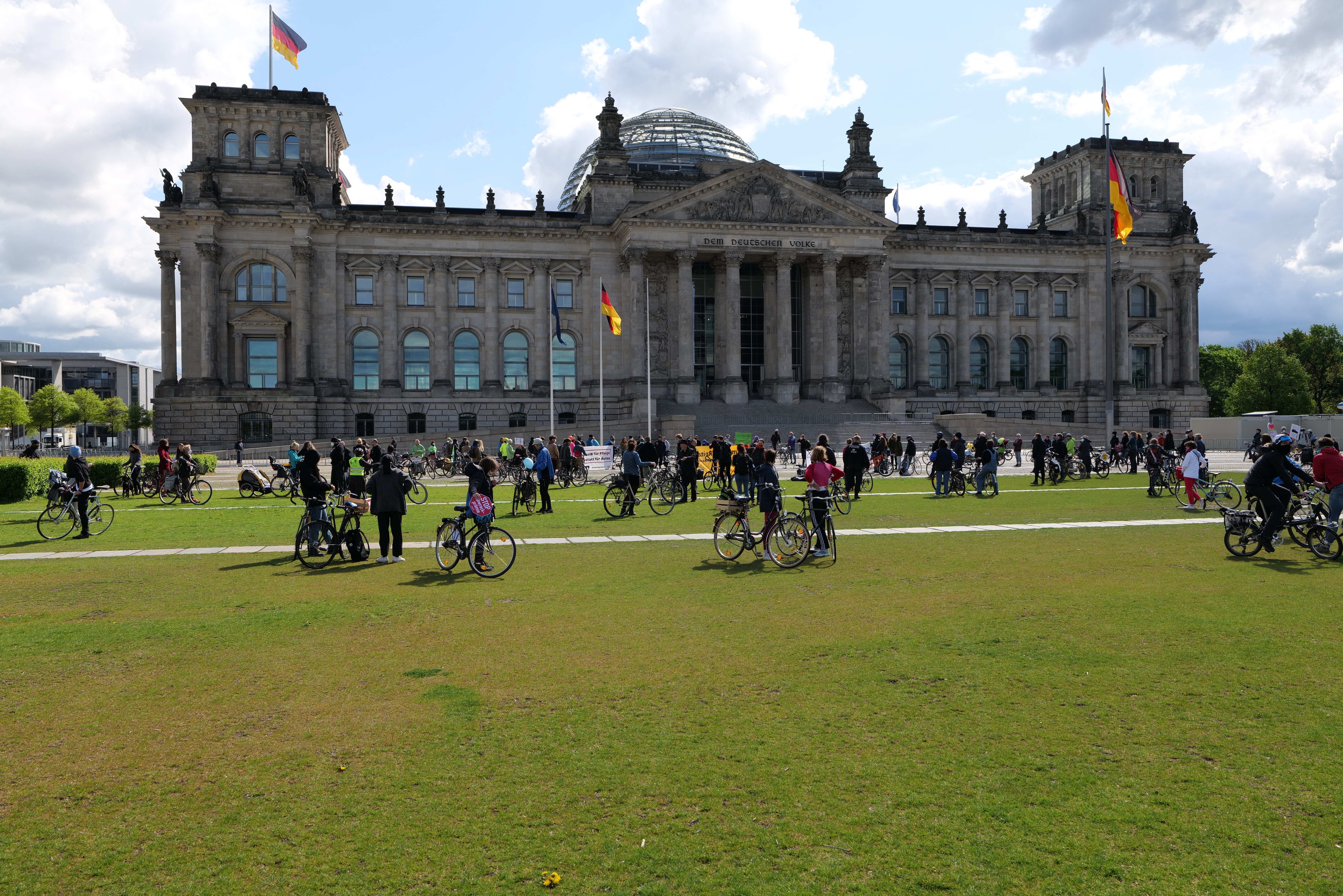 Menschen auf Fahrrädern vor dem Reichstaggebäude in Berlin, Deutschland, mit Bäumen, Pflanzen und einem bewölkten Himmel im Hintergrund.
