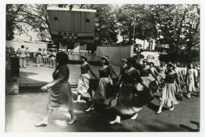 Eine Gruppe von Frauen tanzt auf der Straße, umgeben von einer Menge, einem Lautsprecher, einem Fahrzeug, einer Tafel mit Text, einem Fahnenmast, Gebäuden und einem bewölkten Himmel.
