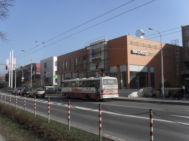 Ein Bus fährt eine Straße mit hohen Gebäuden, Straßenlaternen und Laternenmasten, Autos auf der Straße, Fußgängern auf dem Gehweg und einem klaren blauen Himmel entlang.