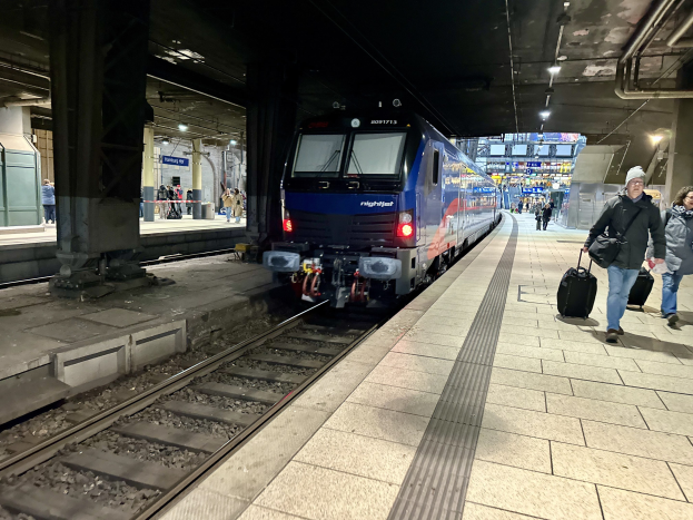 Ein Zug, der nachts in einem hell erleuchteten Bahnhof eintrifft, mit Passagieren auf dem Bahnsteig, die Taschen tragen.
