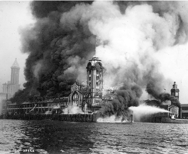 Schwarz-weißes Foto eines großen, von Flammen umloderten Gebäudes mit Rauch, der daraus aufsteigt, einem Gewässer im Vordergrund und anderen Gebäuden und Himmel im Hintergrund.